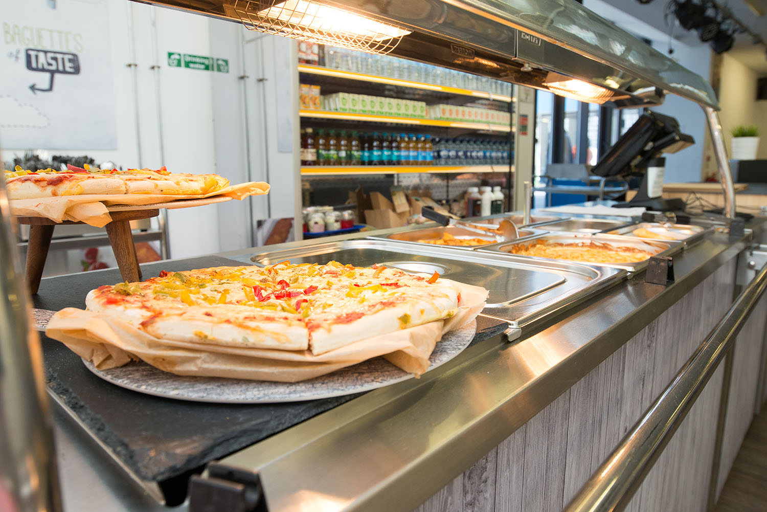 Commercial kitchen serving counter displaying traybake cakes with colourful sprinkles and additional baked goods beneath a heated serving canopy.
Click here to access the week one menu